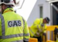 A man in a hi-vis jacket with his back to the camera. On his back is the word GAS. In the background are two people, also in hi-viz jackets, working
