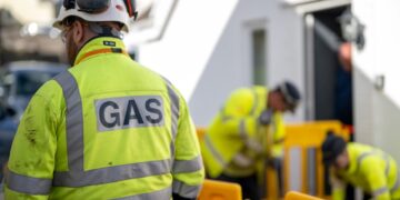 A man in a hi-vis jacket with his back to the camera. On his back is the word GAS. In the background are two people, also in hi-viz jackets, working