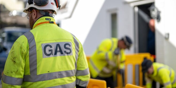 A man in a hi-vis jacket with his back to the camera. On his back is the word GAS. In the background are two people, also in hi-viz jackets, working