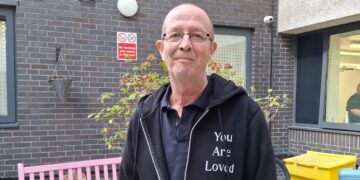 Tim Swinburn looking at the camera. He is outdoors and there is a pink bench behind him