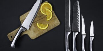 Some kitchen knives on a black background. One knife is on a chopping board with some chopped fruit next to it