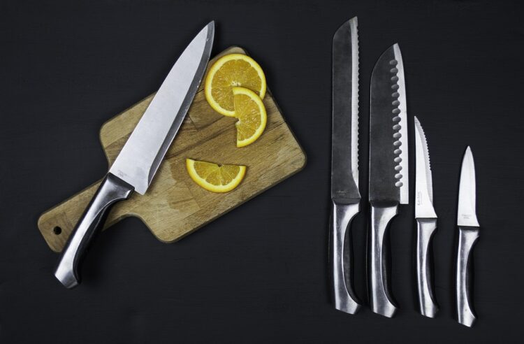 Some kitchen knives on a black background. One knife is on a chopping board with some chopped fruit next to it